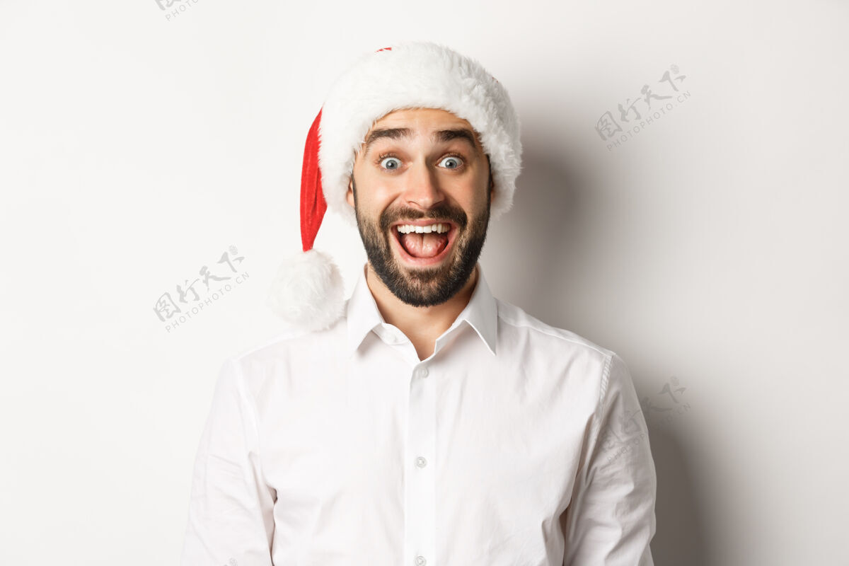 肖像戴着圣诞帽 满脸胡须的快乐男人的特写镜头 看起来很惊讶 正在庆祝圣诞节 站在白色背景上男人黑发休闲