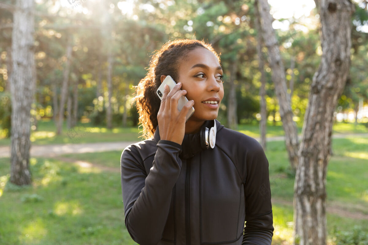 年轻一个20多岁的漂亮女人穿着黑色运动服 戴着耳机 一边在绿色公园里玩手机黑发锻炼户外
