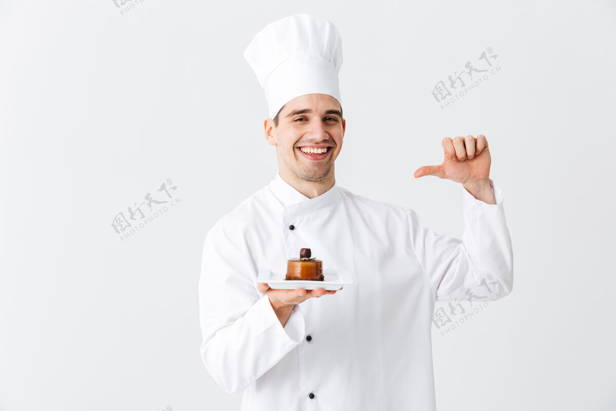 烹饪开朗的厨师穿着制服 在白色的墙上展示一个盘子上的糕点甜点酋长面包师