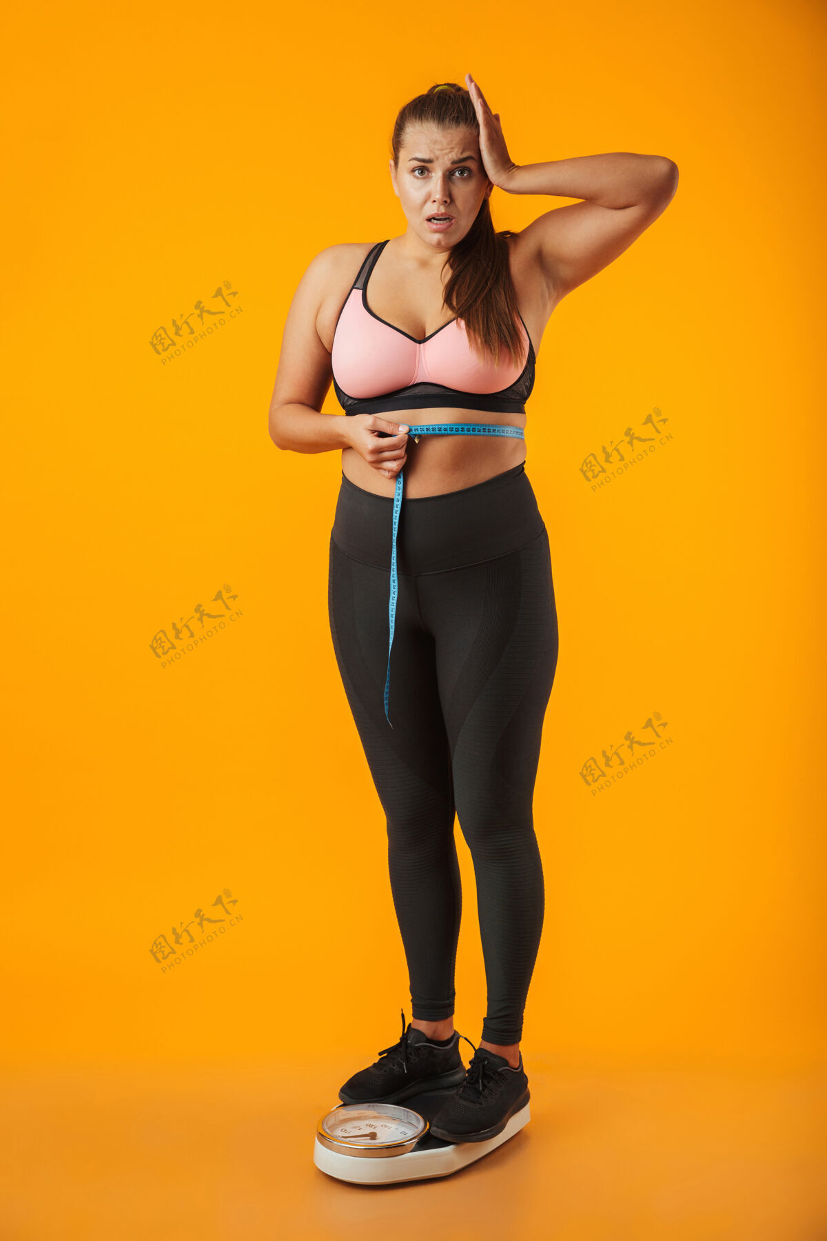 女人一个心烦意乱的超重的年轻女子穿着运动服站在黄色墙壁上 腰间拿着卷尺的全长肖像体重情绪超重