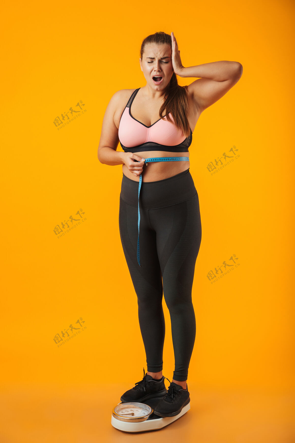 表情一个心烦意乱的超重的年轻女子穿着运动服站在黄色墙壁上 腰间拿着卷尺的全长肖像惊讶身体成就