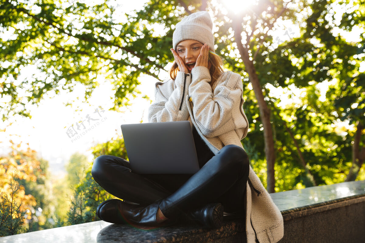 学生美丽的年轻女子身着秋衣 戴着秋帽 坐在户外 用笔记本电脑 庆祝成功职业类型漂亮
