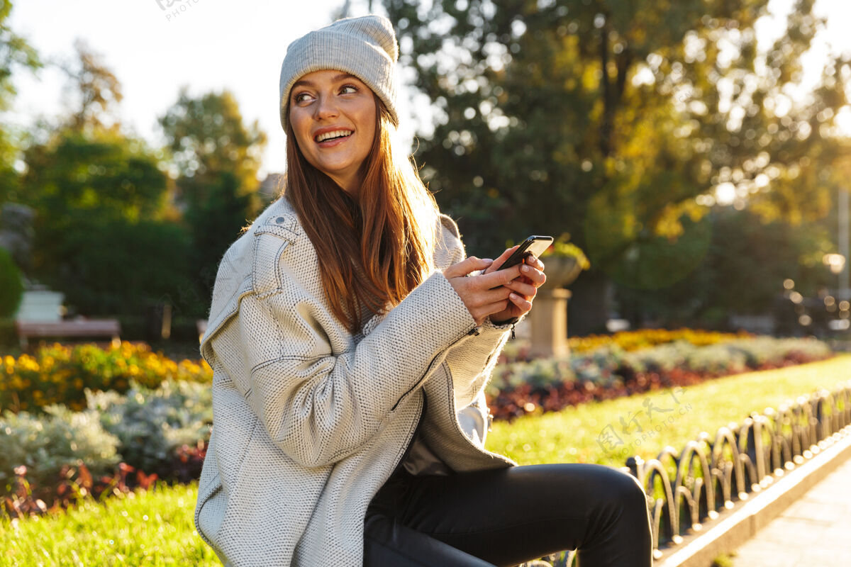 移动美丽的年轻女子穿着秋大衣 戴着秋帽坐在户外的长凳上 用手机早晨举行魅力