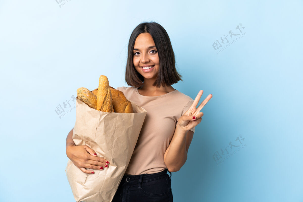 烹饪一个年轻的拉丁女人在买面包 脸上挂着蓝色的微笑和胜利的标志面包人成功