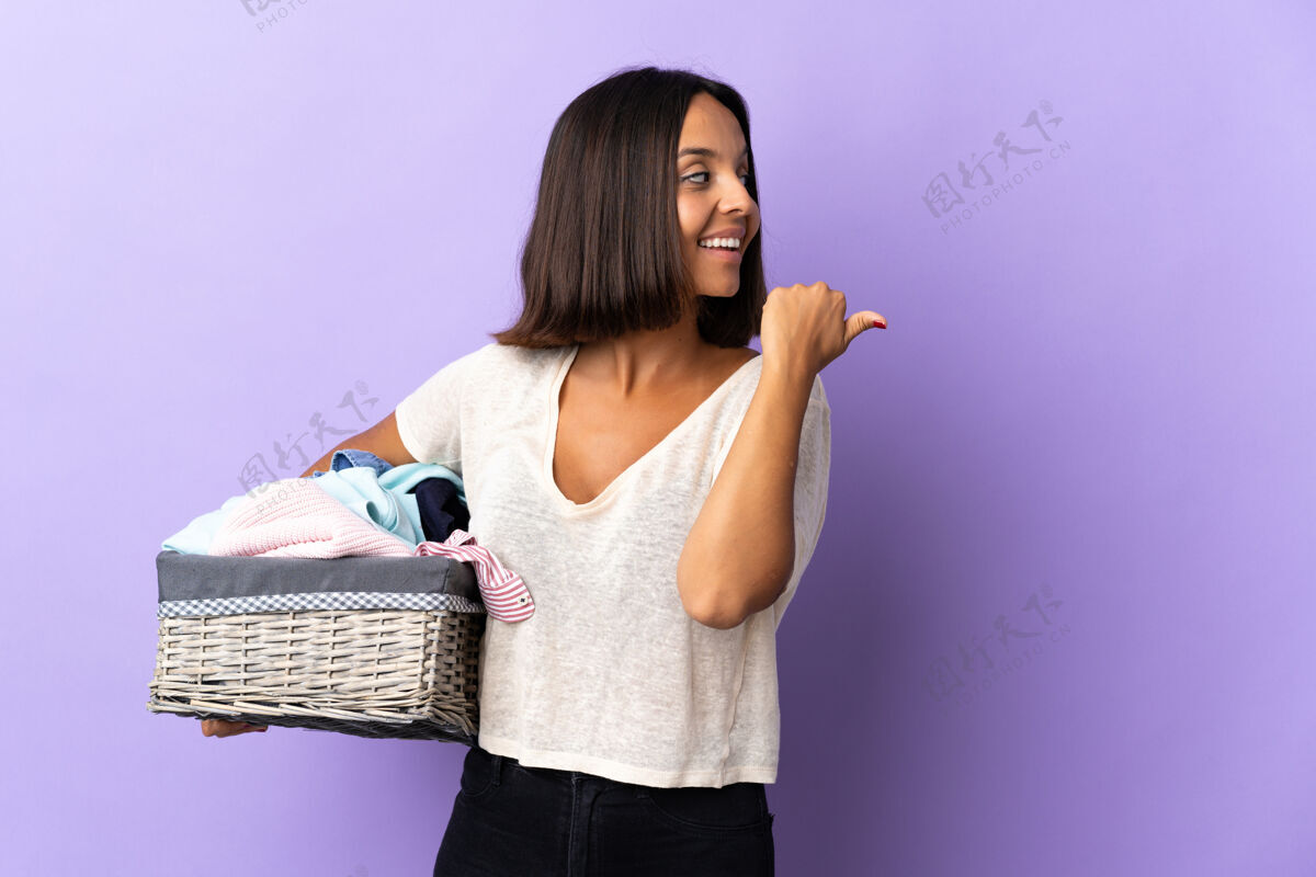 人年轻的拉丁女子拿着一个衣篮孤立地站在紫色的边上指着礼品洗衣服赛车衣服