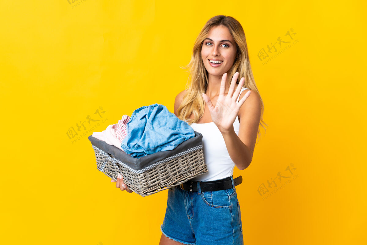 脏年轻的金发乌拉圭女人拿着一个衣篮 孤立地站在黄色的地上 用手指数着五个女人年轻洗衣机