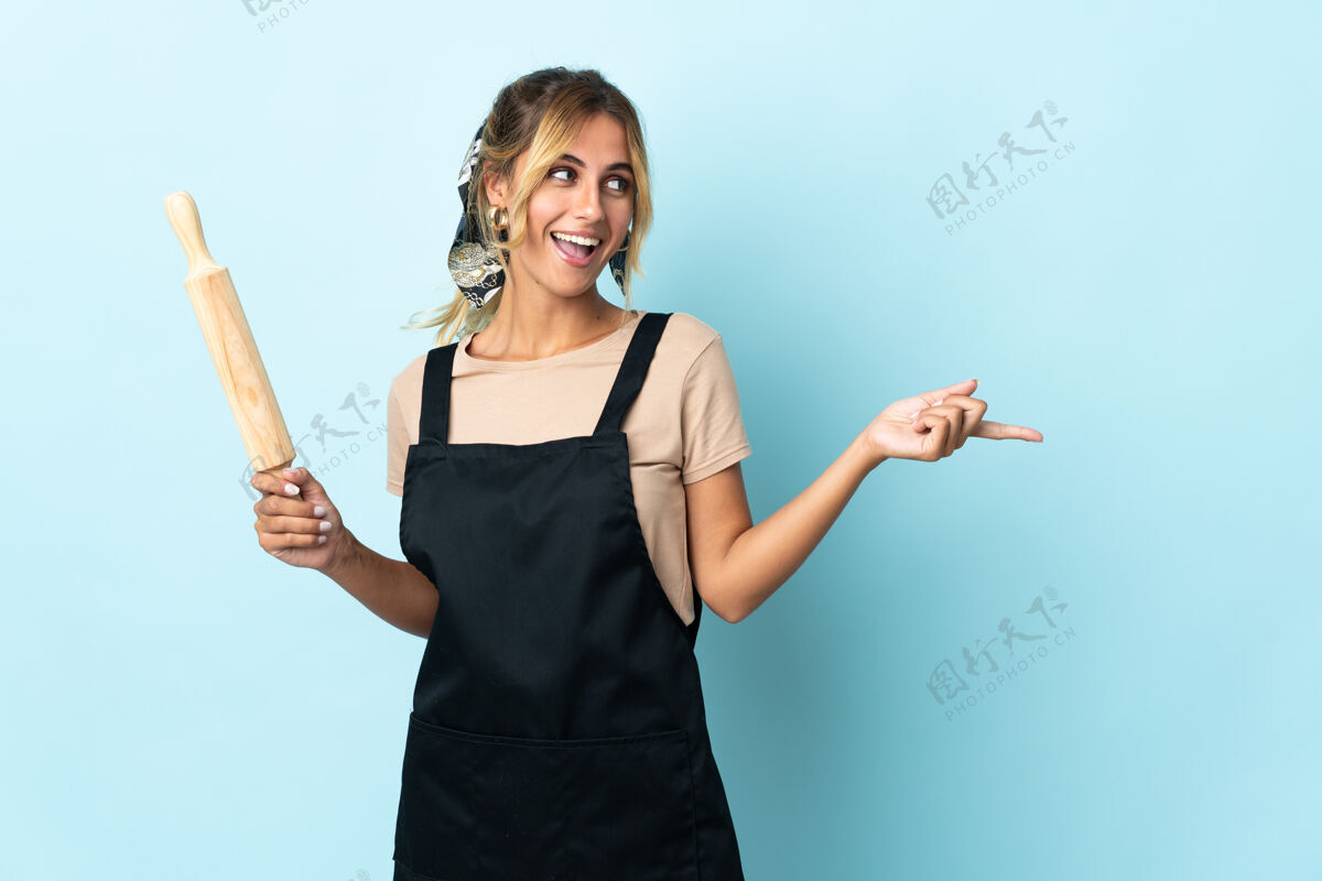 围裙年轻的金发乌拉圭厨娘孤立地用蓝色手指指着旁边 并介绍一个产品女面包房烹饪