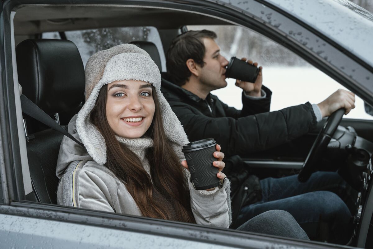 公路旅行侧视图笑脸夫妇在车上享受一个温暖的饮料 而在公路旅行汽车女性雪