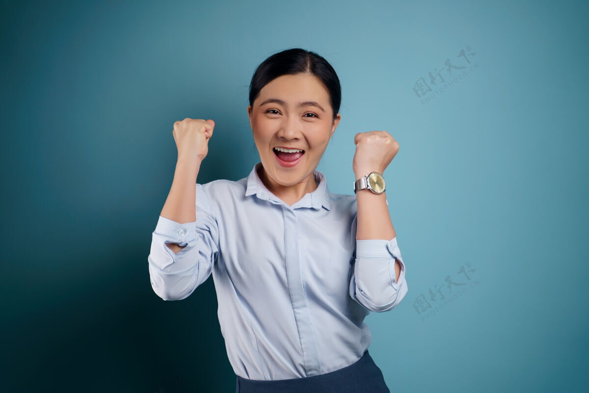积极的情绪亚洲女人快乐自信的站着 展示她的拳头 做一个胜利的手势 孤立在蓝色上表达友好思考