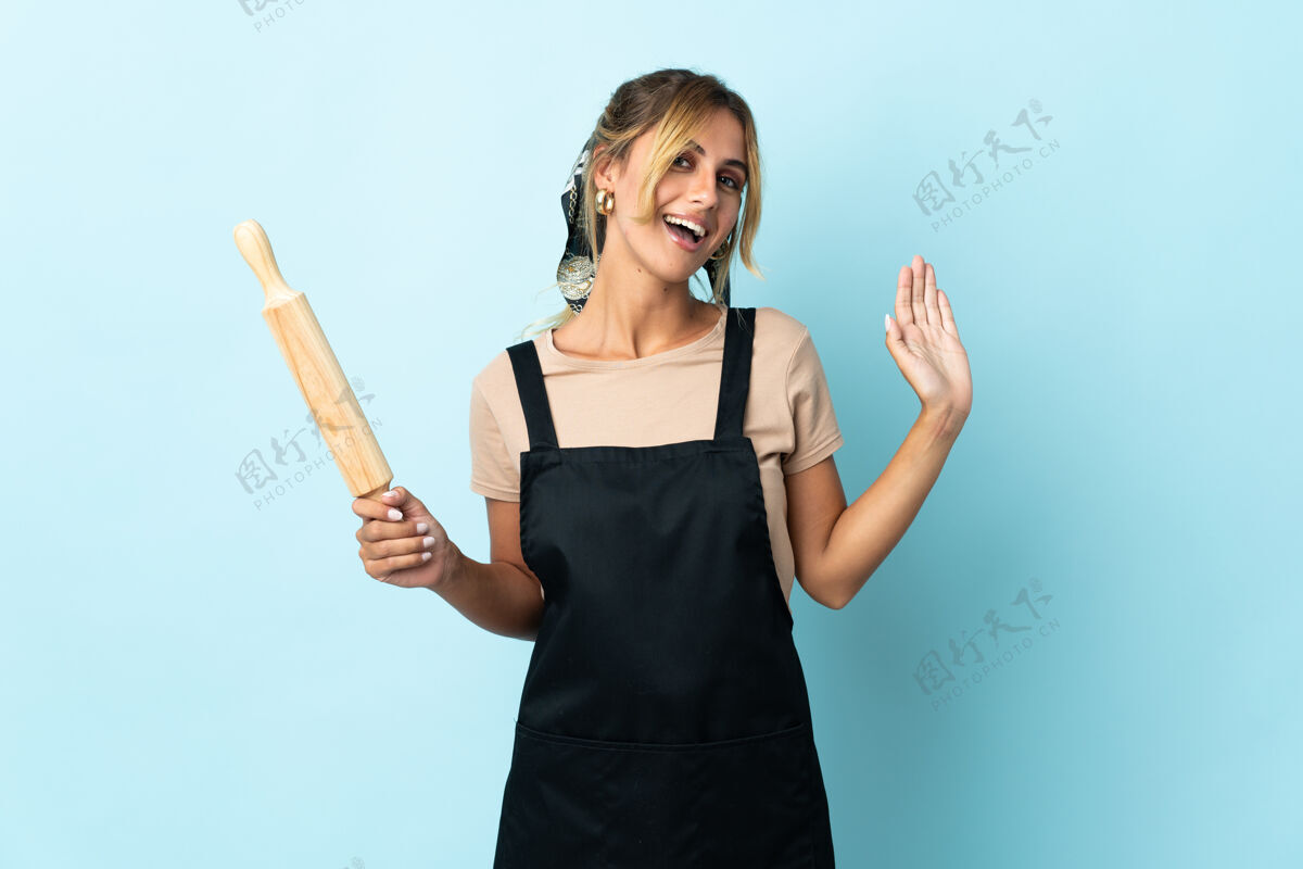 女人年轻的金发乌拉圭厨娘孤立在蓝色的行礼手与快乐的表情厨房面包师工作