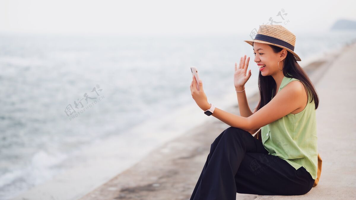 微笑在海边的海滩上 一位亚洲旅行者正在使用智能手机在线视频通话帽子旅游快乐