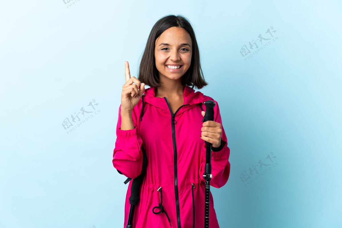 手指年轻的拉丁女人背着背包和蓝色的登山竿提出了一个好主意女人走路远足者