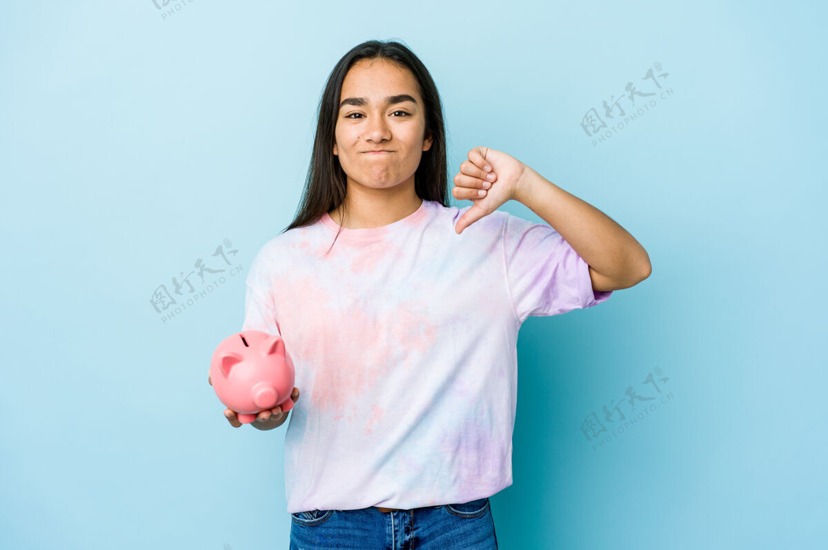 拇指一位年轻的亚洲女士拿着一个粉红色的银行 站在隔离墙上 用拇指做着一个讨厌的手势不同意概念储蓄女孩不喜欢