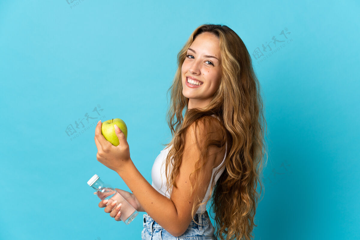 苹果年轻的金发女人拿着一个苹果和一瓶水被隔离在蓝色的天空中人食物女人