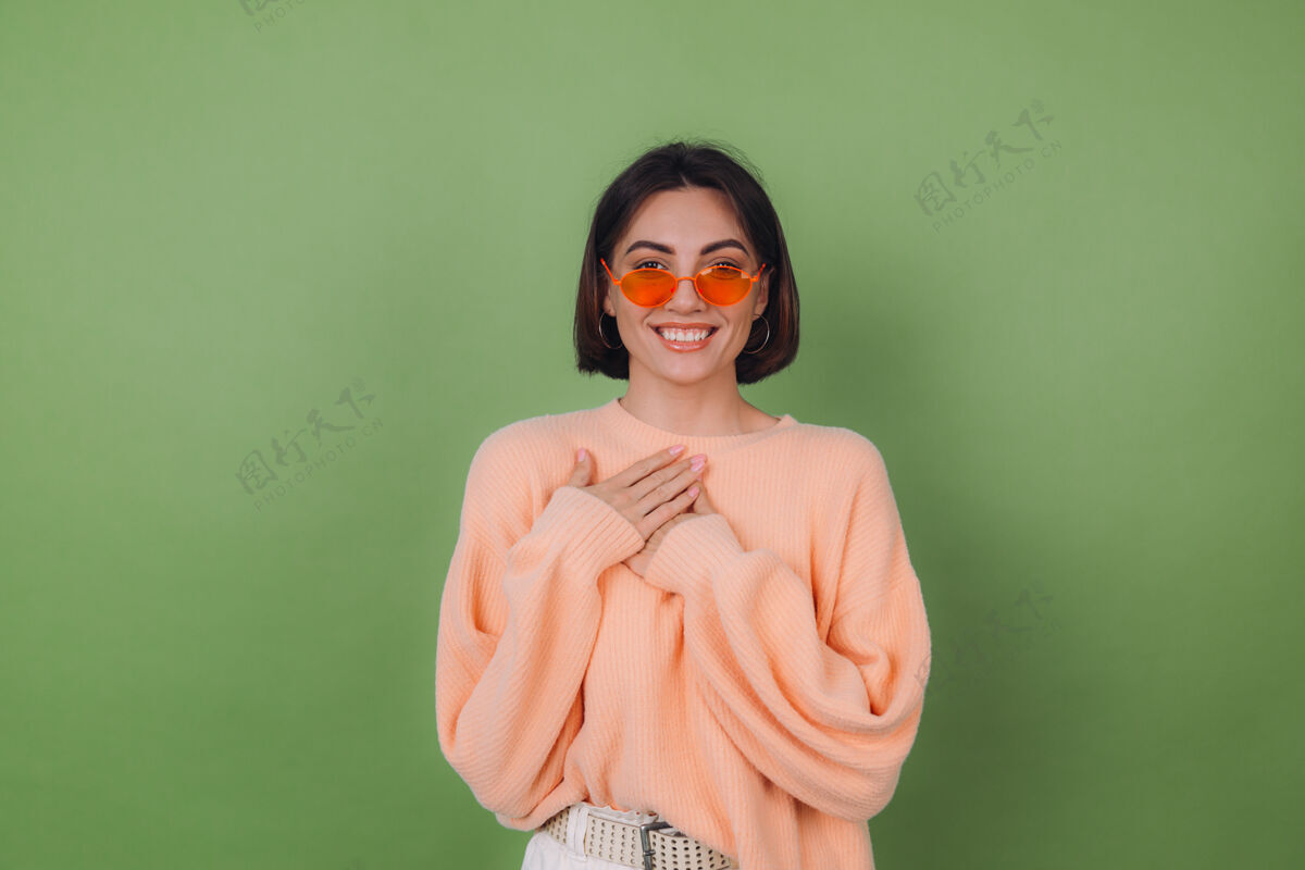 人年轻时尚的女士穿着休闲桃色毛衣 戴着橘色眼镜 隔着绿橄榄色的墙正面双手合十放在胸前 心复制空间感激女人黑发