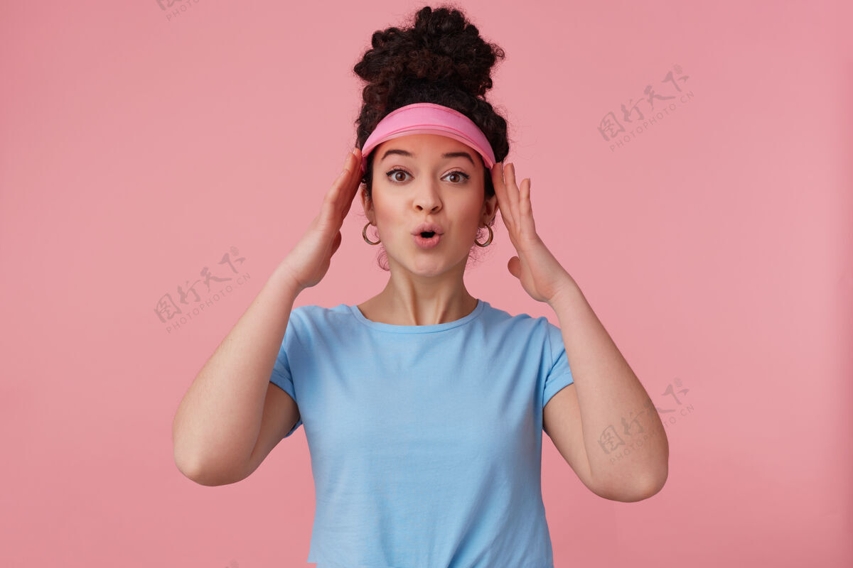 女士惊艳的女人 美丽的女孩 深色卷发 戴着粉红色的帽舌 耳环和蓝色的t恤 化妆 抚摸着她的头化妆品遮阳板T恤