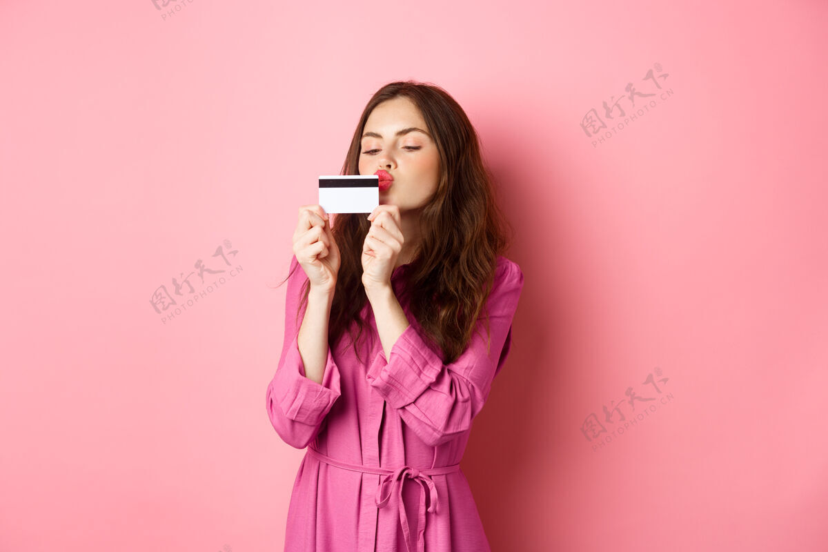 乐观年轻漂亮的女人亲吻她的信用卡 去购物 在商店里浪费钱 站在粉红色的墙上候选人手势女孩