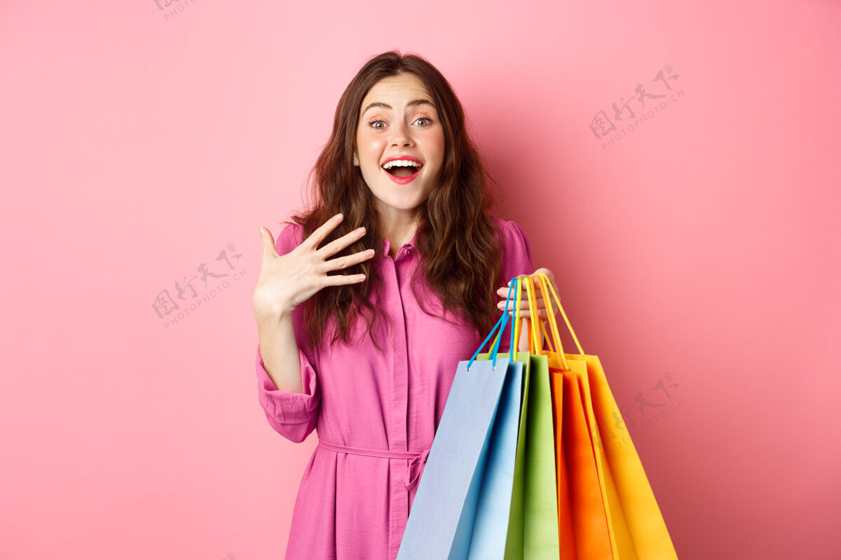女孩年轻的女士手拿着粉红色的购物袋 站在墙上 惊讶地喘息着女人休闲年轻