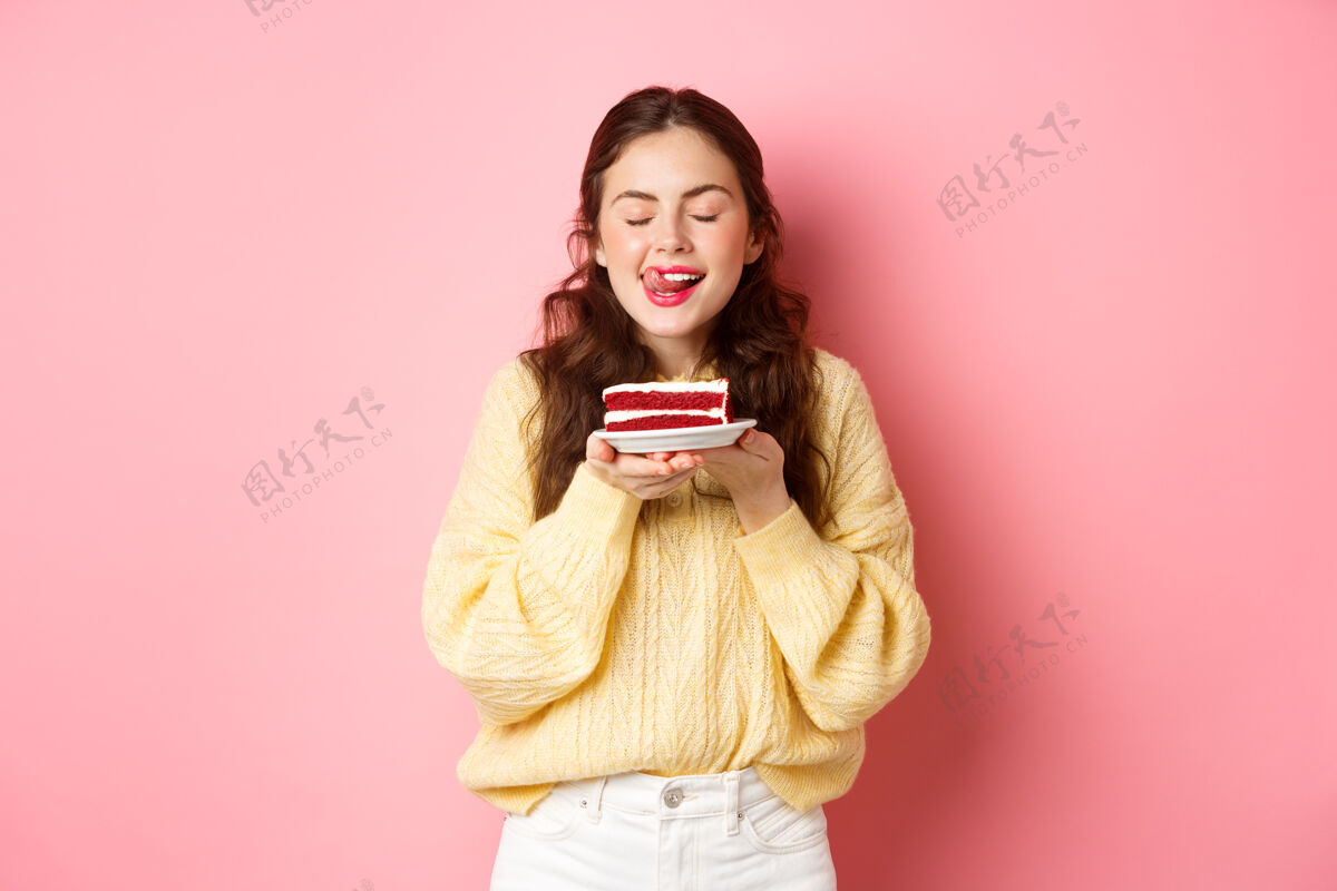 乐观女人拿着美味的蛋糕 闭着眼睛 梦幻般地舔着嘴唇 想吃美味的甜点 站在粉红色的墙上休闲快乐时尚