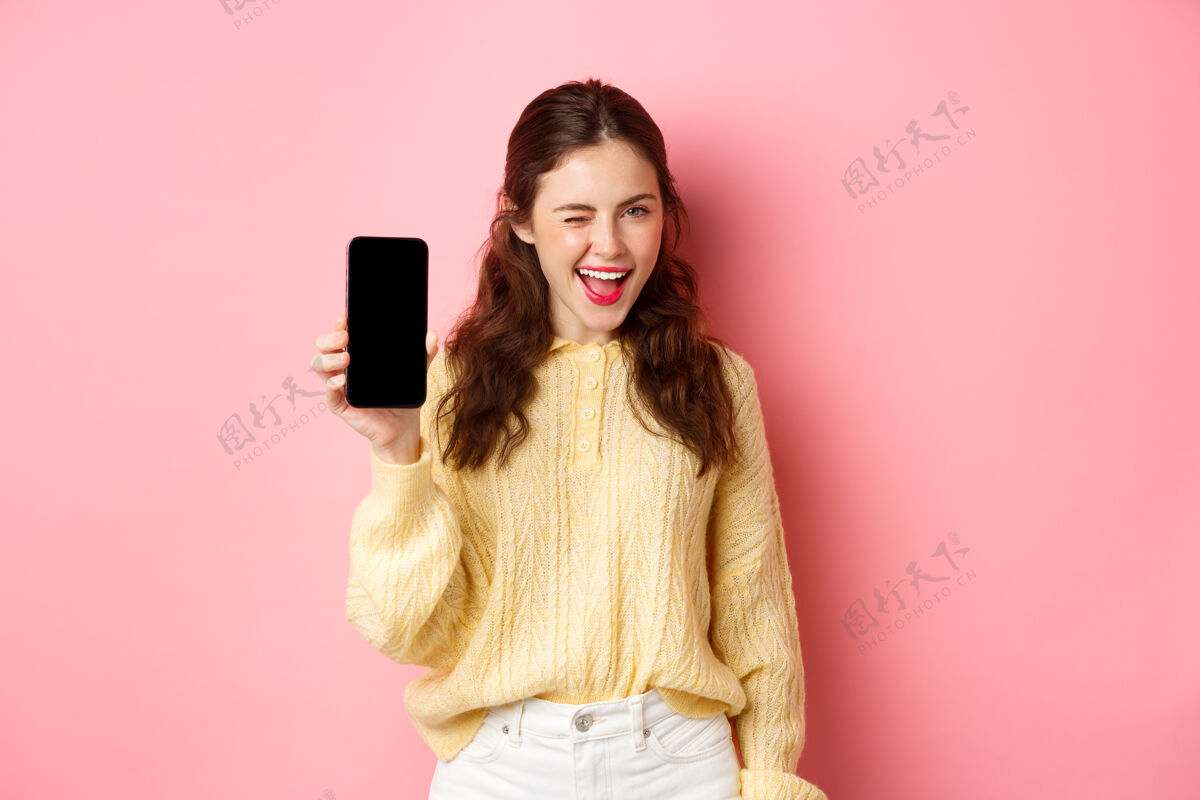 年轻厚颜无耻的漂亮女孩 给你看智能手机屏幕 眨眼微笑 推荐手机 站在粉红色的墙上手机时尚水平