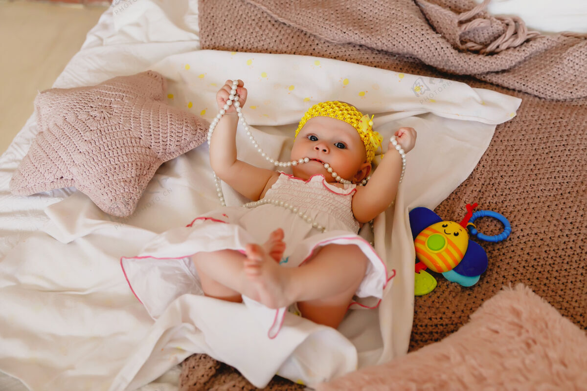玩耍可爱快乐的蓝眼睛6个月大的白衣女孩在床上玩珍珠珠女孩婴儿表情
