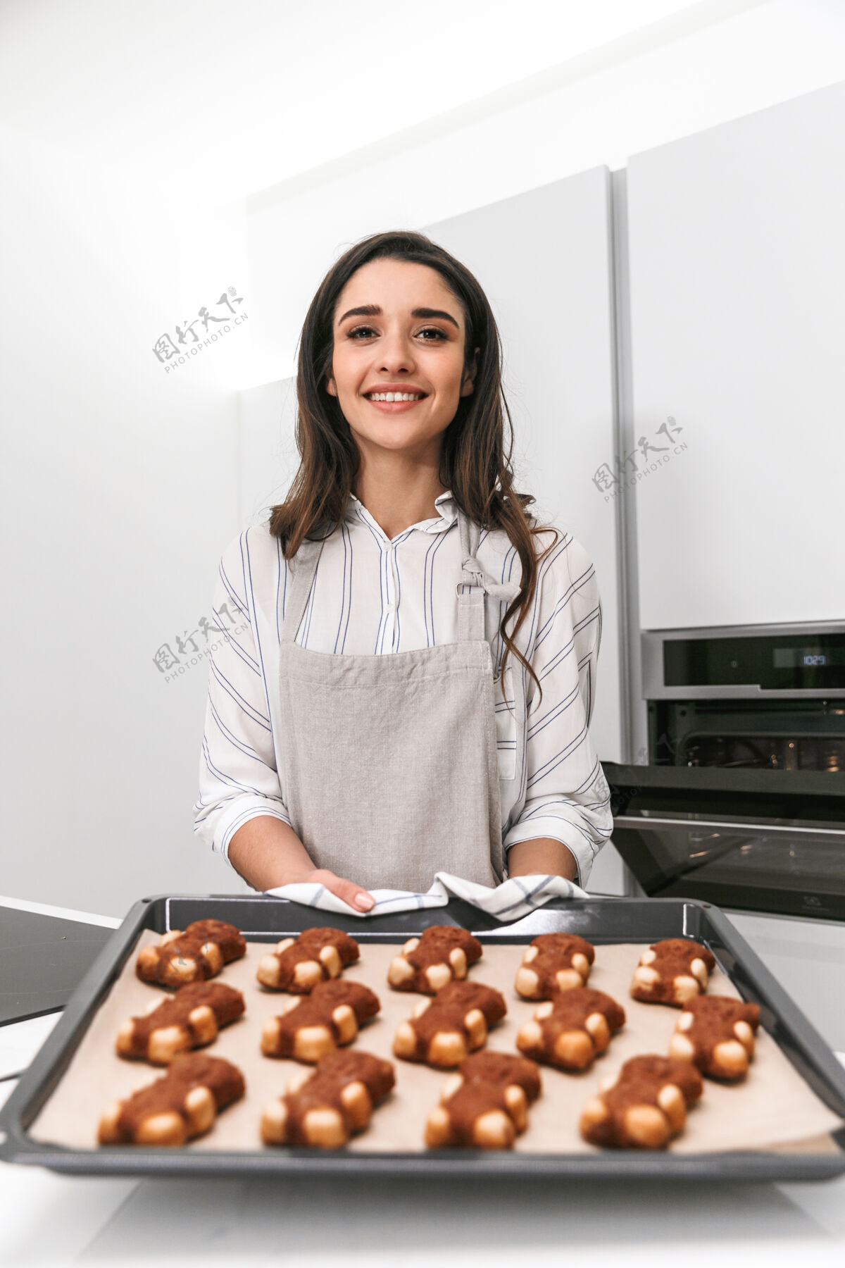 围裙站在厨房里用托盘做美味饼干的迷人年轻女子年轻热厨师