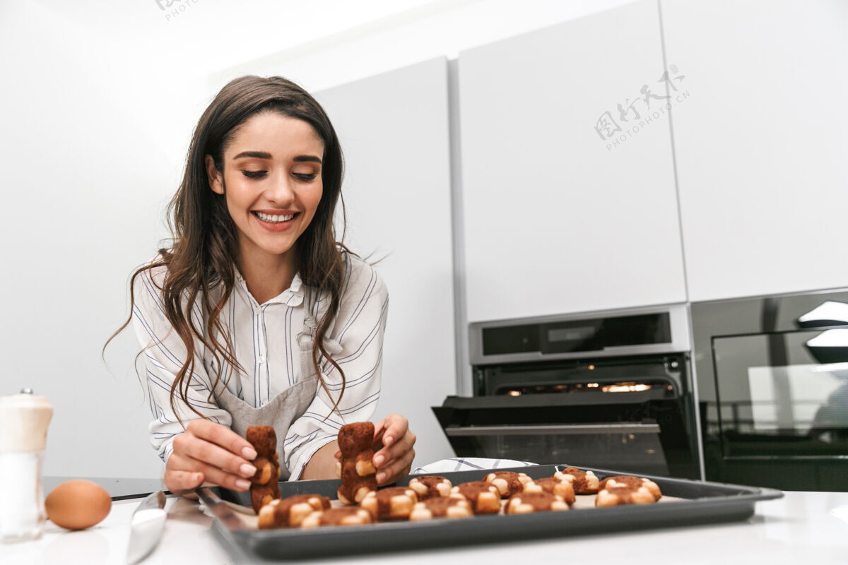 蛋糕站在厨房里用托盘做美味饼干的迷人年轻女子热食物围裙
