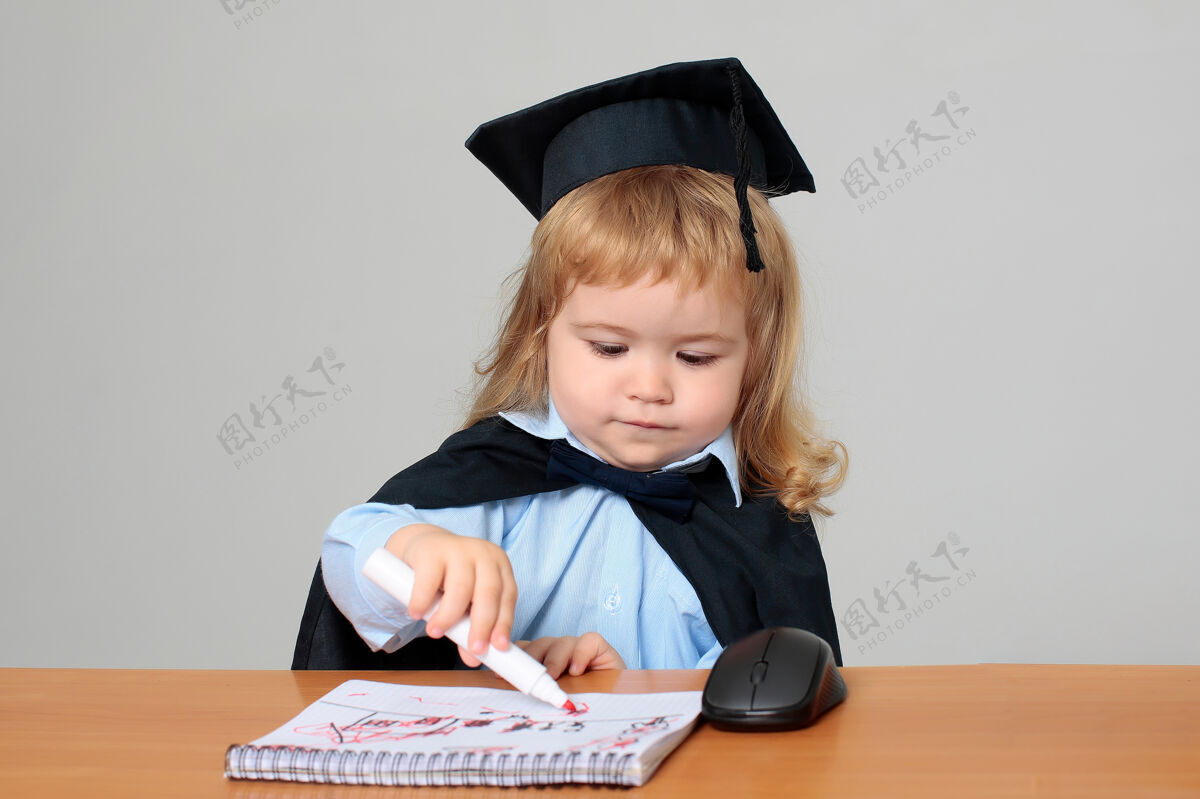 马克穿着毕业礼服 戴着毕业帽的学生宝宝在课桌旁用马克笔在笔记本上画画图画教学灯泡