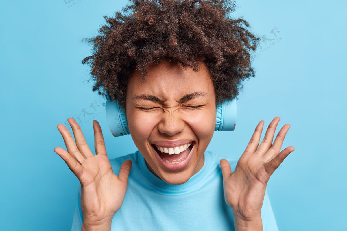 享受喜出望外的美国黑人妇女举手高呼高兴地闭上眼睛对可怕的新闻作出反应戴着无线耳机隔着蓝色的墙壁快乐的概念位置快乐穿着