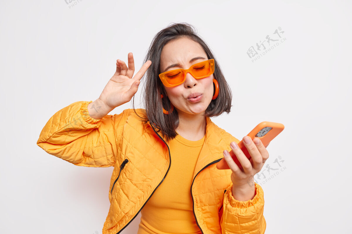 享受喜气洋洋的黑发亚裔女子随着喜爱的歌曲翩翩起舞随着音乐的节奏手持现代智能手机戴着时尚的橙色太阳镜时尚的夹克表达着室内模特们的快乐和喜悦人类青少年穿着