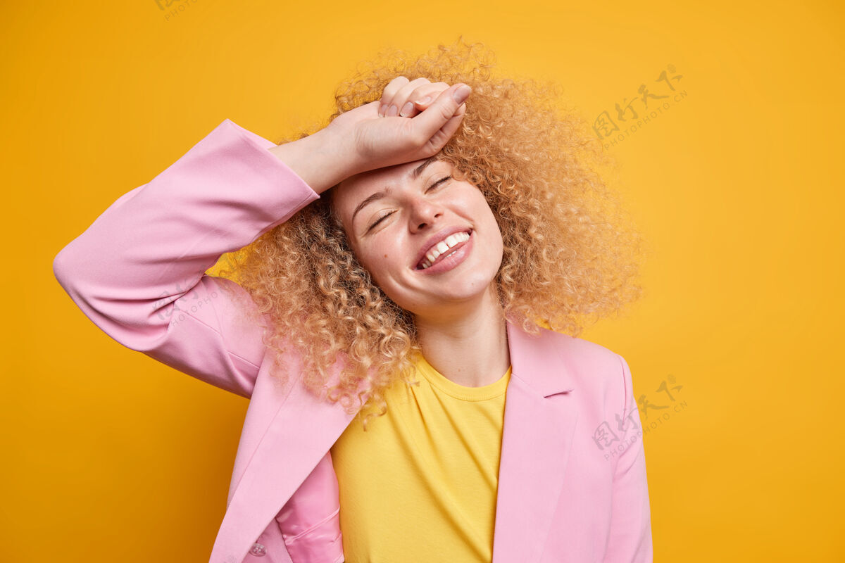 额头自然卷发的无忧无虑的女人微笑着 积极地闭上眼睛 手放在额头上 穿着正式的衣服 在鲜艳的黄色墙壁上表达幸福幸福的概念时尚快乐人