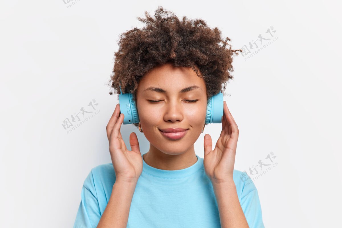 人放松的卷发女士合影闭上眼睛欣赏音乐手戴耳机音质好穿着休闲蓝色t恤隔着白墙生活方式概念肖像黑发卷发