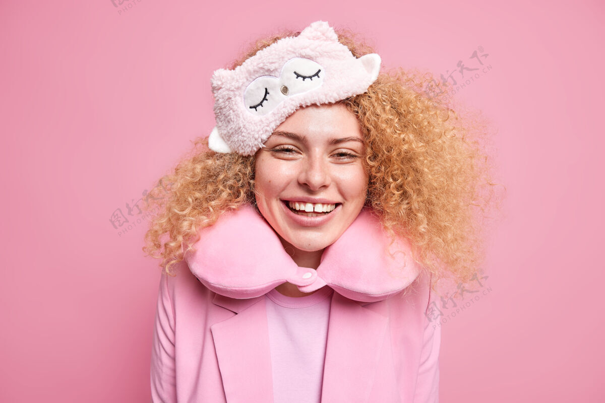 人开朗的年轻女模特 卷曲浓密的头发 笑容满面 心情愉快地醒来 戴着睡眠面罩和颈枕 舒适地休息 快乐地站在粉红色的墙上早上好枕头衣服室内