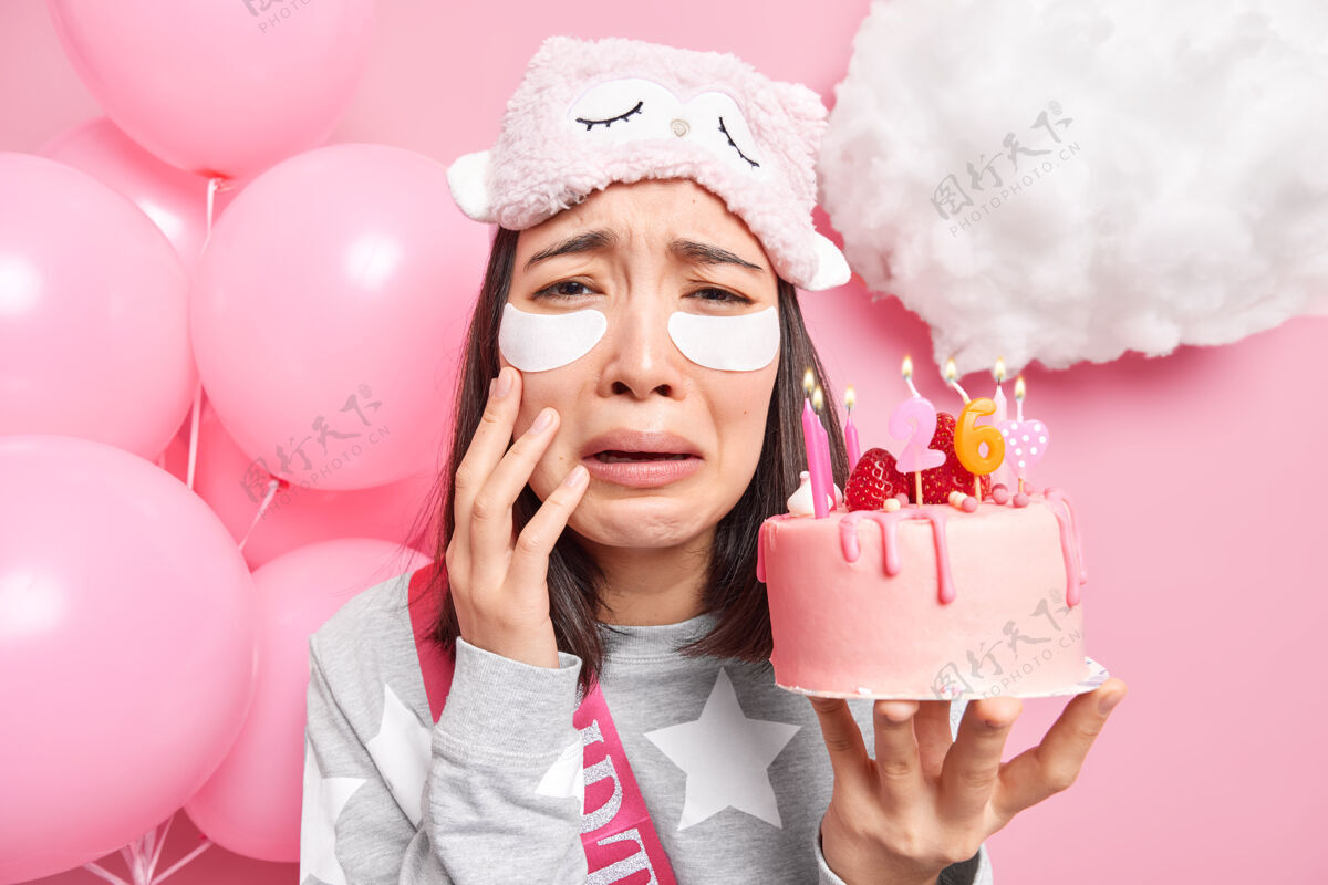 悲伤女人在她生日的时候感到孤单 在眼睛下面涂上美容贴 看起来很美 拿着蛋糕点着蜡烛房子睡衣气球