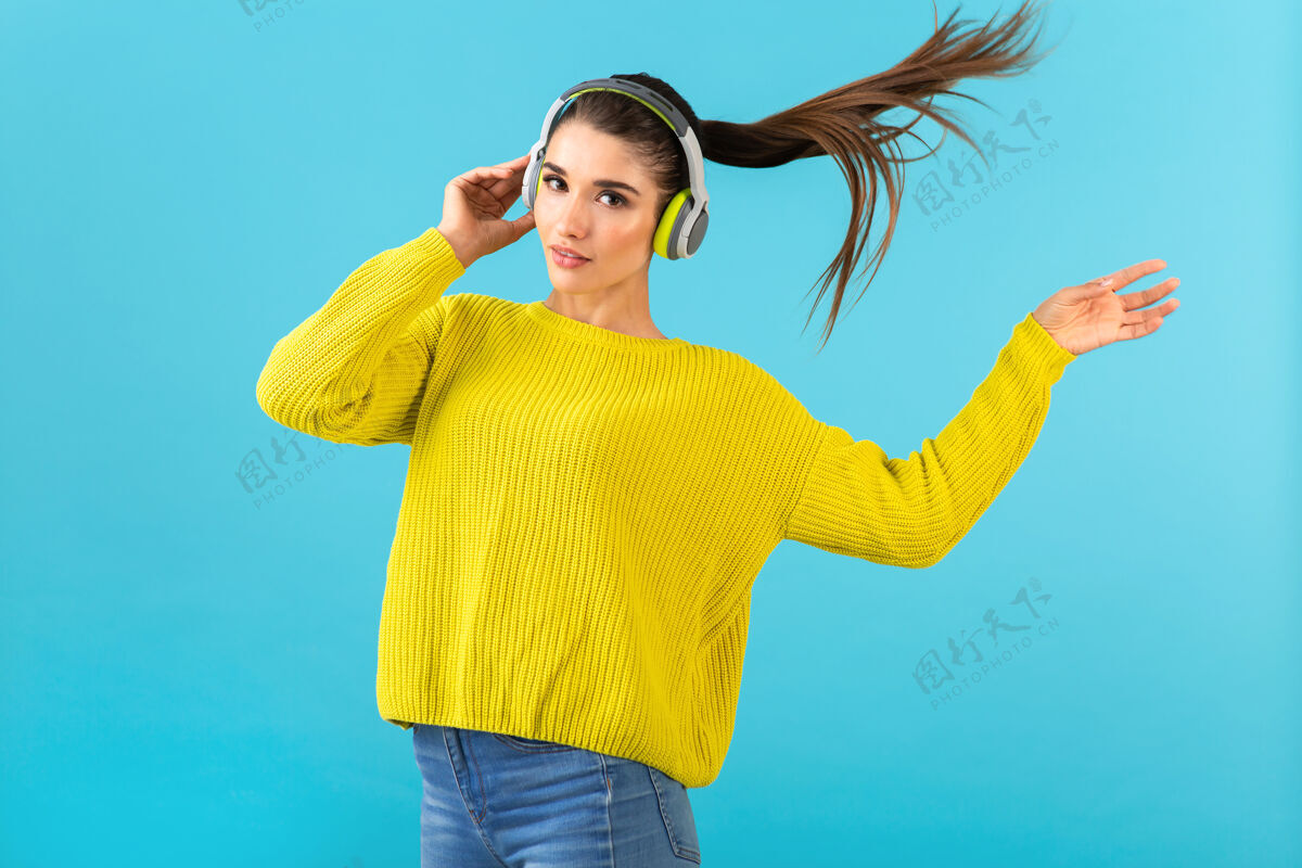 时髦迷人时尚的年轻女子戴着无线耳机听音乐快乐地穿着黄色针织毛衣五颜六色的时尚造型摆姿势酷漂亮耳机
