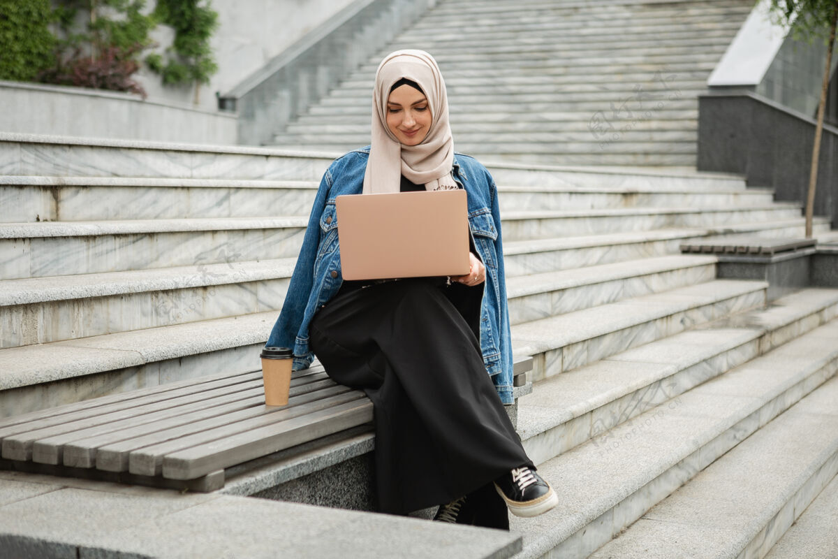 伊斯兰现代时尚的穆斯林妇女戴着头巾 穿着牛仔夹克和黑色长袍坐在城市街道上用笔记本电脑工作穆斯林职业头巾