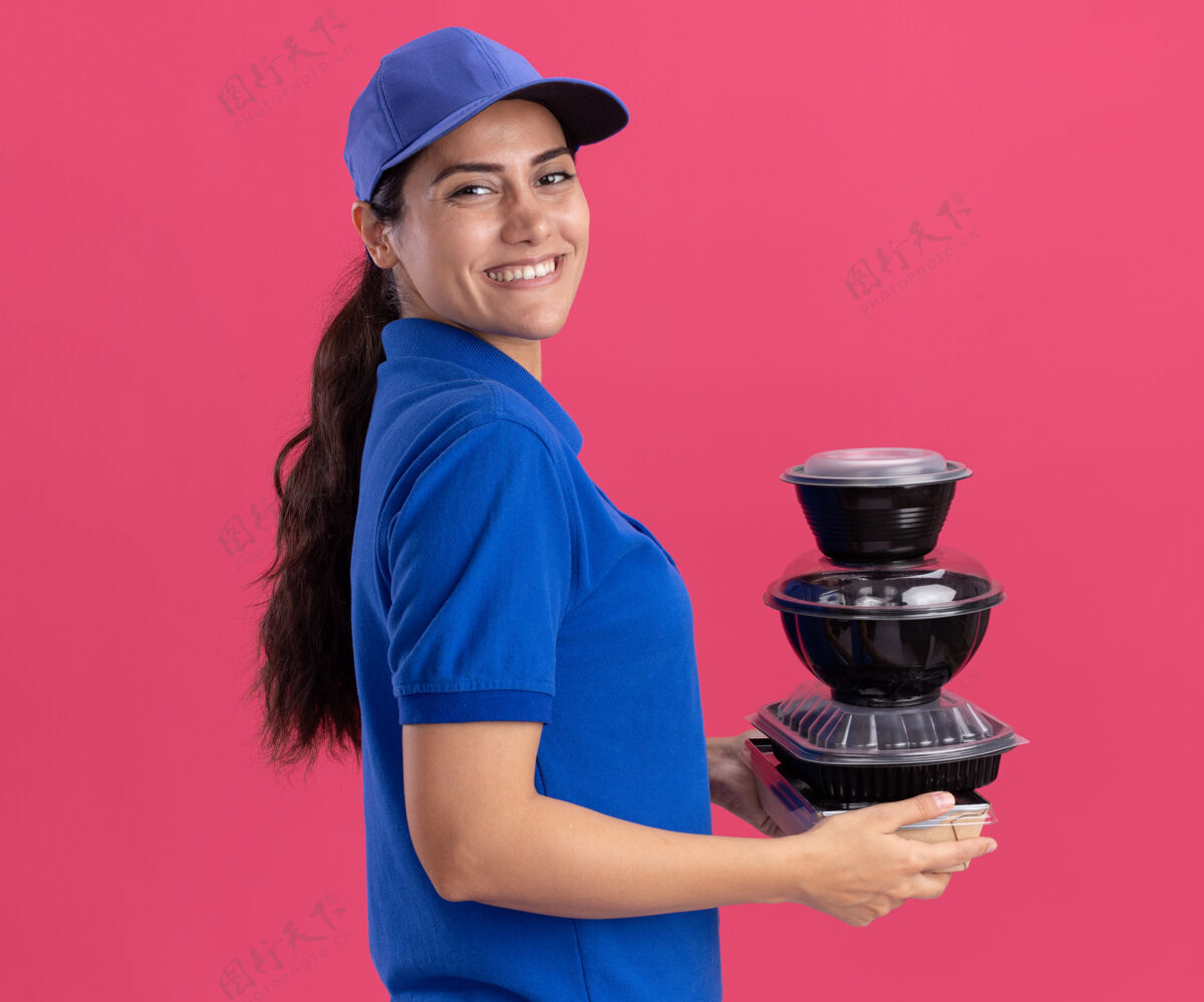 食物微笑着站在剖面图年轻的送货女孩穿着制服 帽子拿着食物容器隔离在粉红色的墙上衣服感觉人