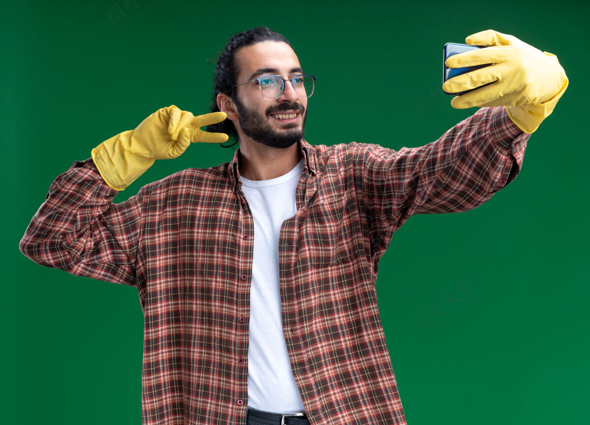 公民微笑着的年轻帅气的清洁工 穿着t恤和手套 在绿色的墙上做一个显示和平姿态的自拍英俊穿人