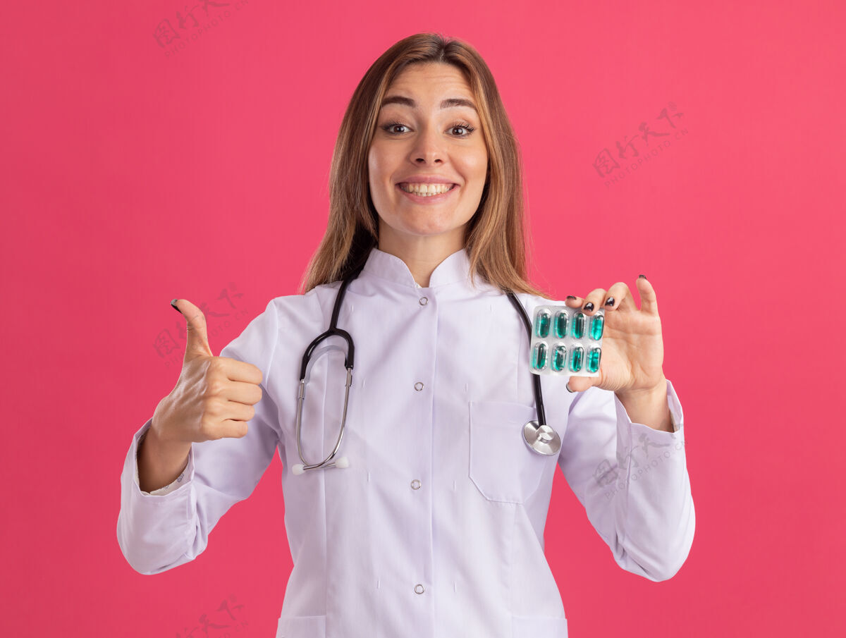 公民面带微笑的年轻女医生穿着医用长袍 手持听诊器的药片 在粉红色的墙上孤立地竖起大拇指年轻人穿着姿势