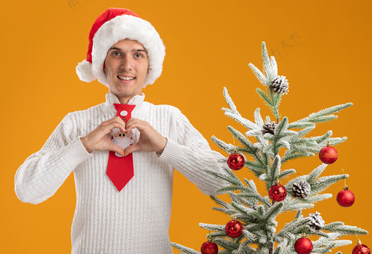 圣诞老人微笑的年轻帅哥戴着圣诞帽 打着圣诞老人的领带 站在装饰好的圣诞树旁做着心形的手势 看起来孤立在橙色的墙上年轻心小伙子