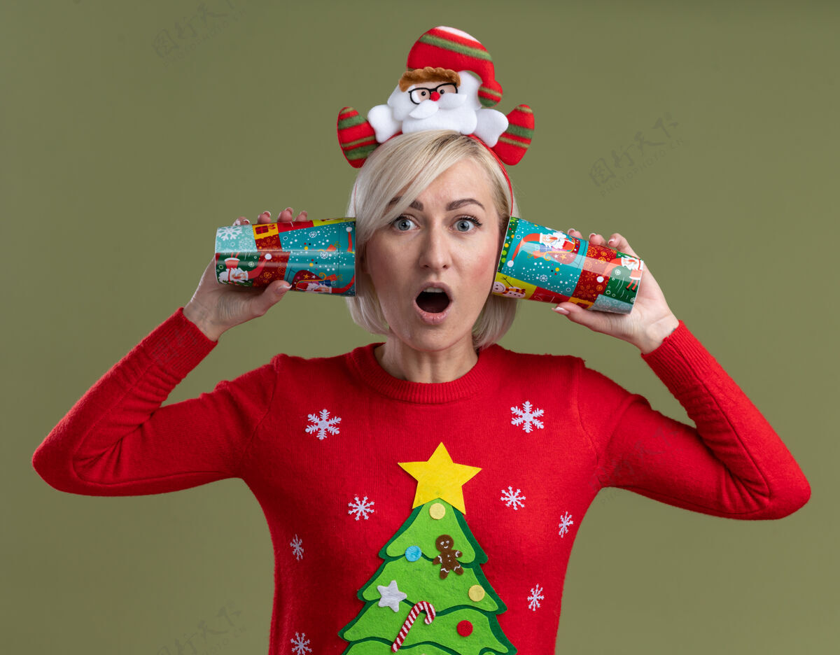 穿惊讶的中年金发女人戴着圣诞老人的头带和圣诞毛衣 手里拿着塑料圣诞杯 耳边听着橄榄绿墙上孤立的对话塑料头带杯子