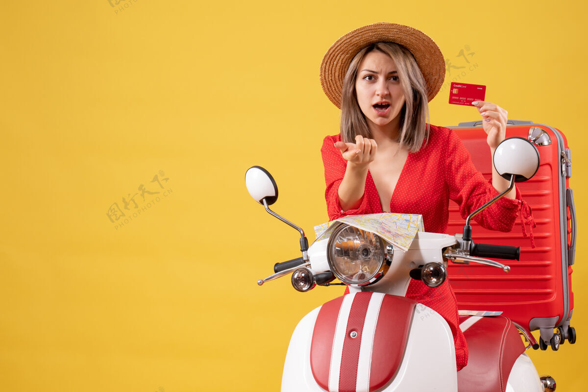 红色前视图穿着红色裙子的年轻女士拿着信用卡指着附近的轻便摩托车行李旅游行李
