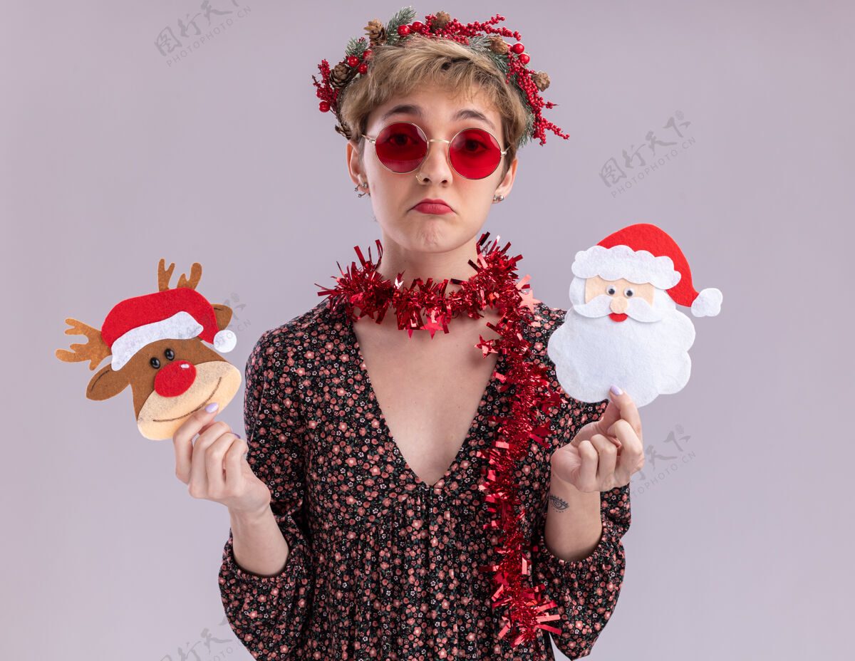 花环无知的年轻漂亮女孩戴着圣诞花环 脖子上戴着金属丝花环 戴着眼镜 把圣诞驯鹿和圣诞老人的纸饰品孤立在白色的墙上头圣诞节年轻