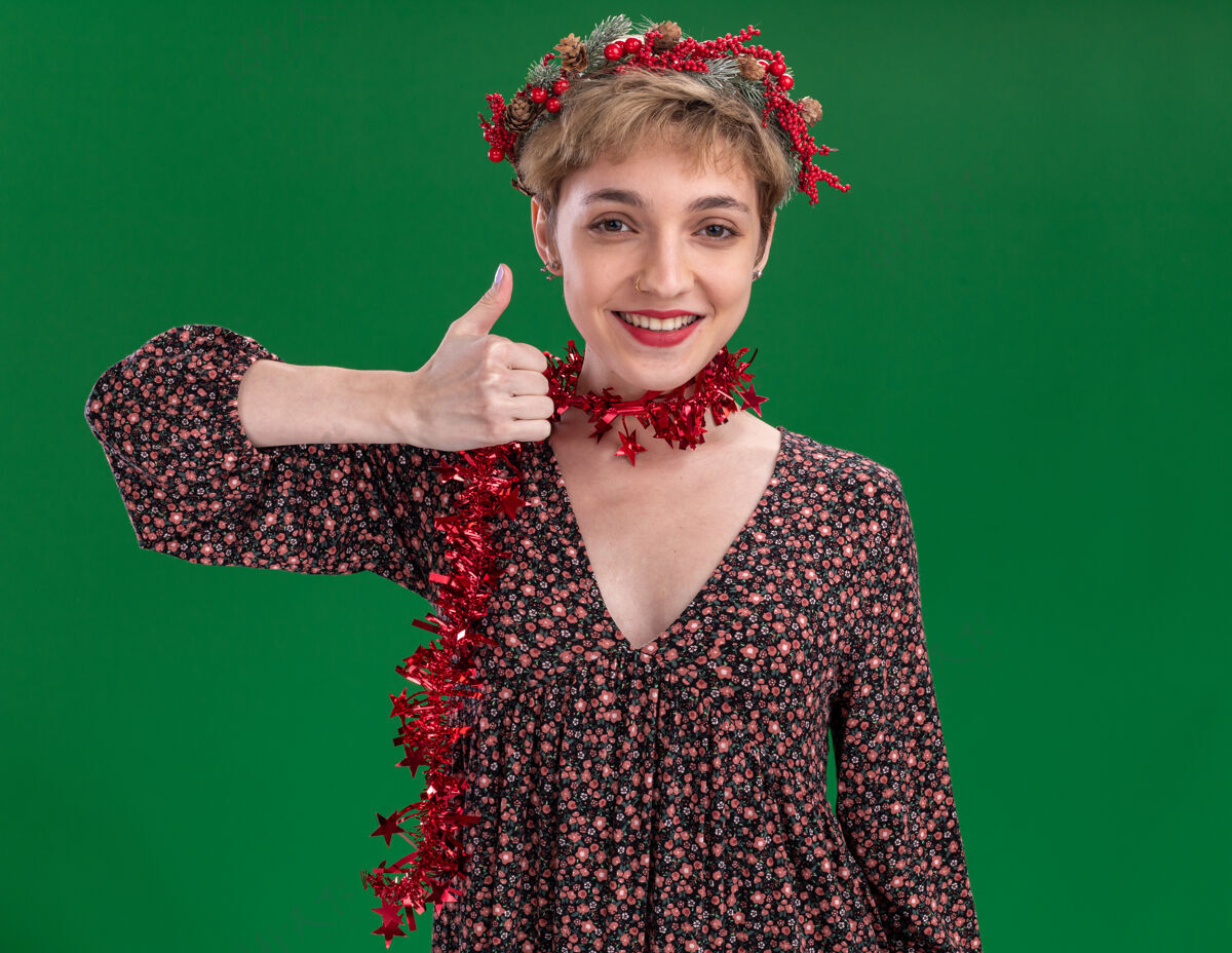 头微笑着的年轻漂亮女孩戴着圣诞花环 脖子上戴着金箔花环 在绿色的墙上孤立地竖起大拇指显示圣诞节漂亮
