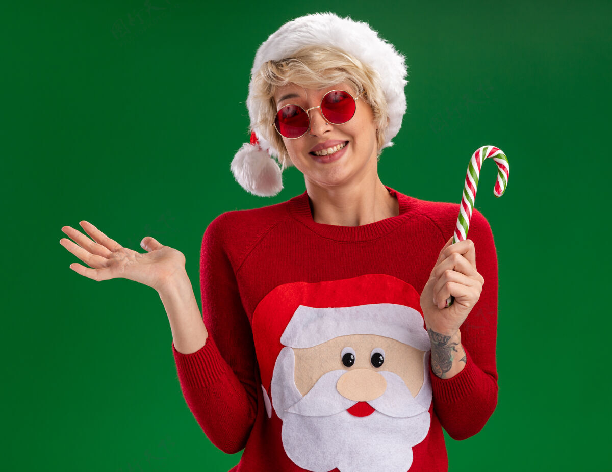 穿微笑的金发女郎戴着圣诞帽和圣诞老人的圣诞毛衣戴着圣诞眼镜拿着圣诞糖果手杖看着空空的手孤立在绿色的墙上圣诞老人秀女人