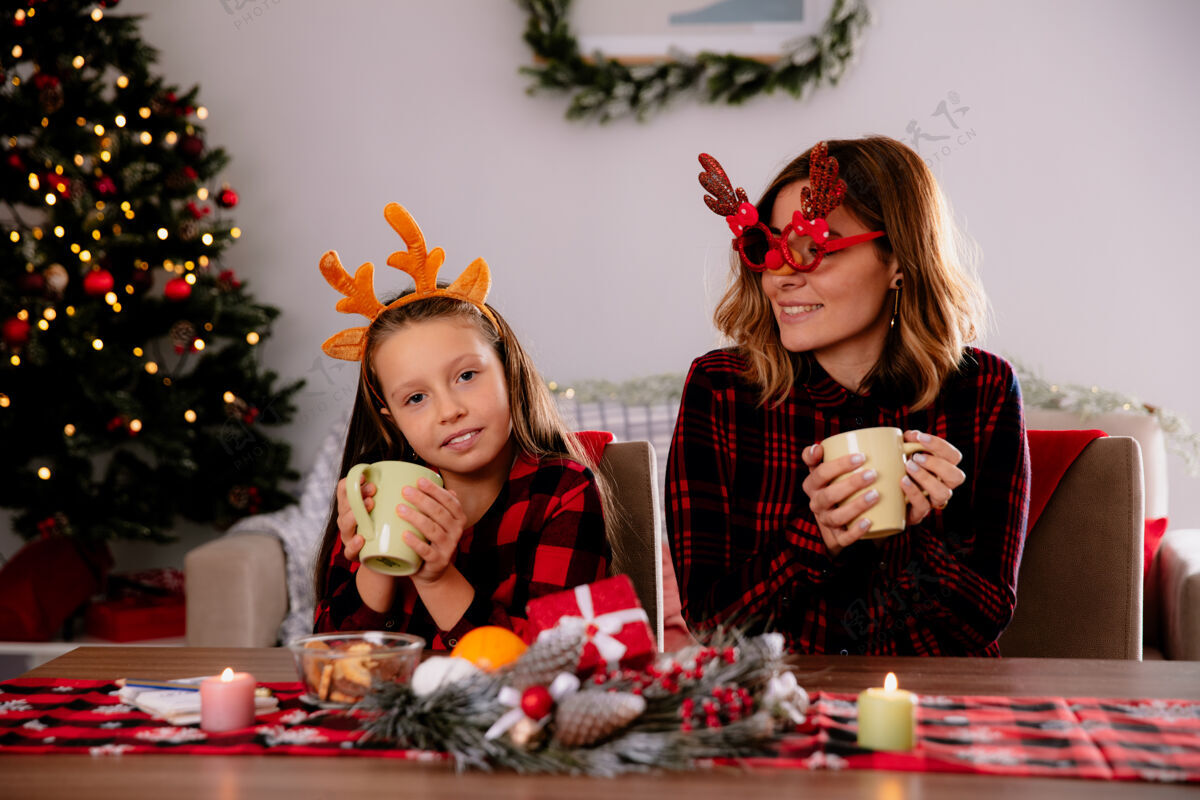 杯子戴着驯鹿眼镜的妈妈高兴地看着拿着杯子坐在桌边的女儿在家里享受圣诞节的时光请享受女儿杯子