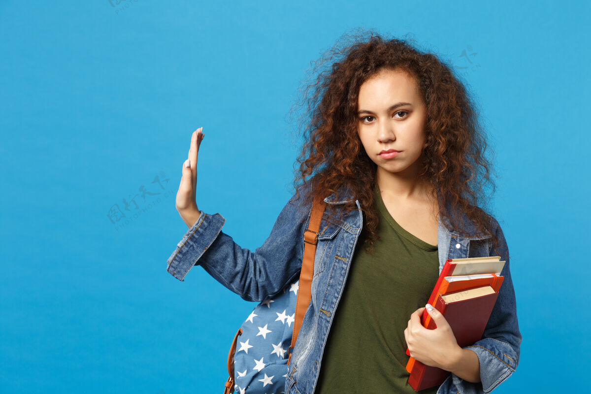 站着穿着牛仔服 背着书包的悲伤的年轻学生把书孤立地放在蓝色的墙上教育阅读包