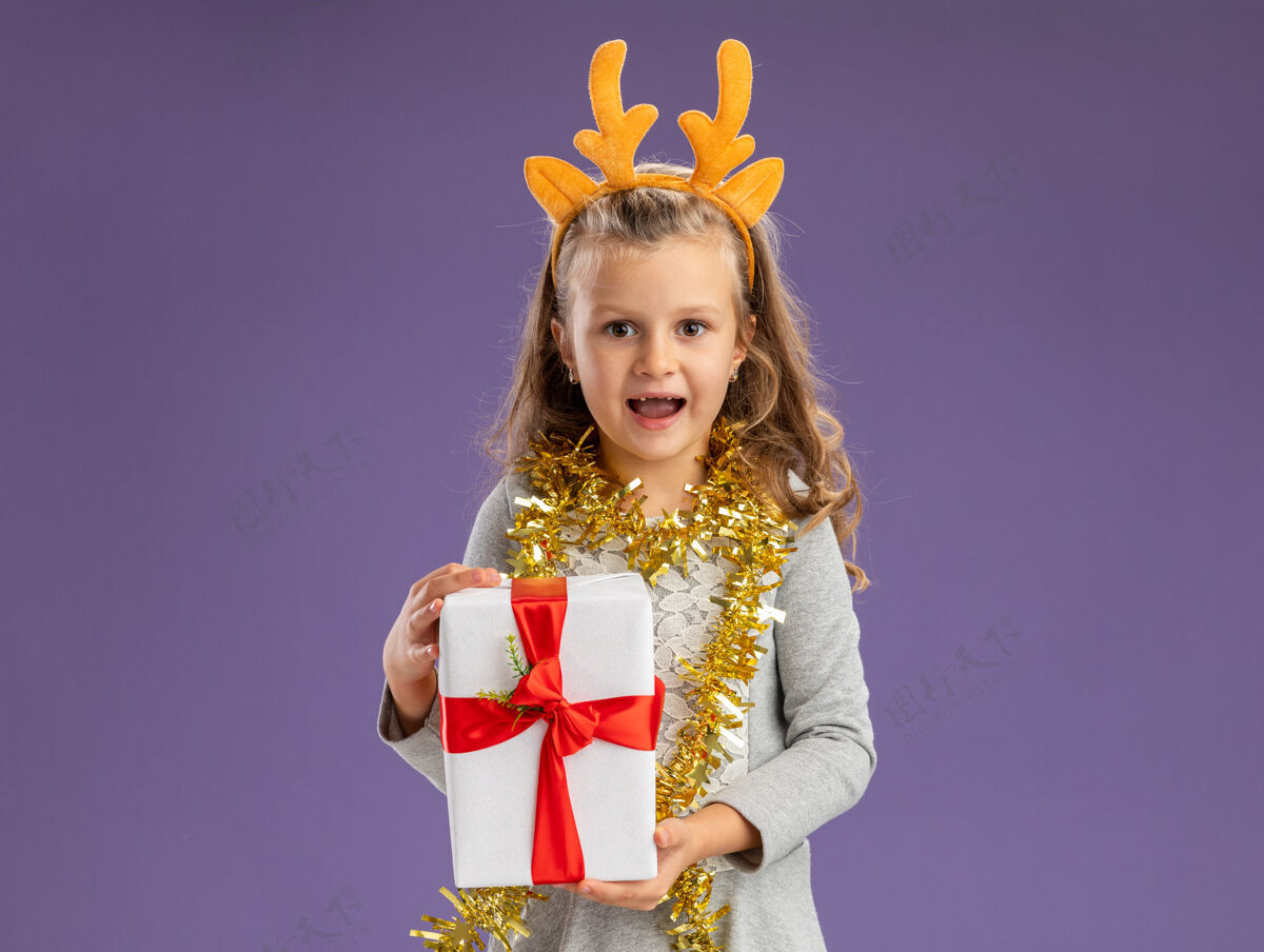女孩微笑的小女孩戴着圣诞发箍 脖子上戴着花环 手里拿着礼品盒 隔离在蓝色的墙上脖子微笑花环