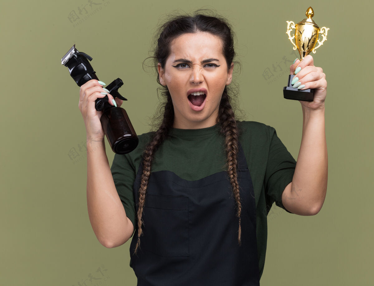 兴奋兴奋的年轻女理发师穿着制服 手里拿着优胜者杯 橄榄绿的墙上孤立着理发器脸杯子人