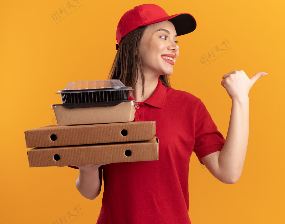 拿着一个穿着制服 面带微笑的漂亮送货员拿着比萨饼盒上的纸质食品包 看着并指着旁边女人纸指点